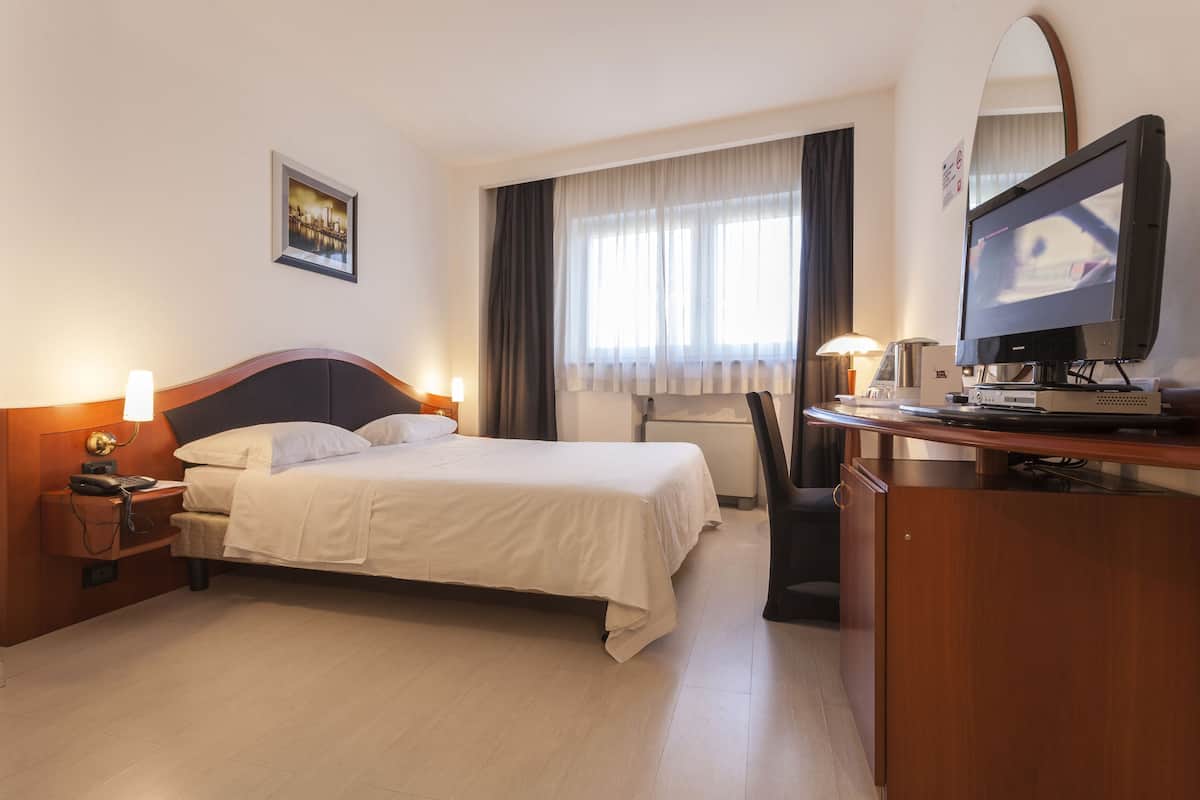 A hotel room with a bed, bedside table, lamp, TV, desk, chair, and a window with curtains.