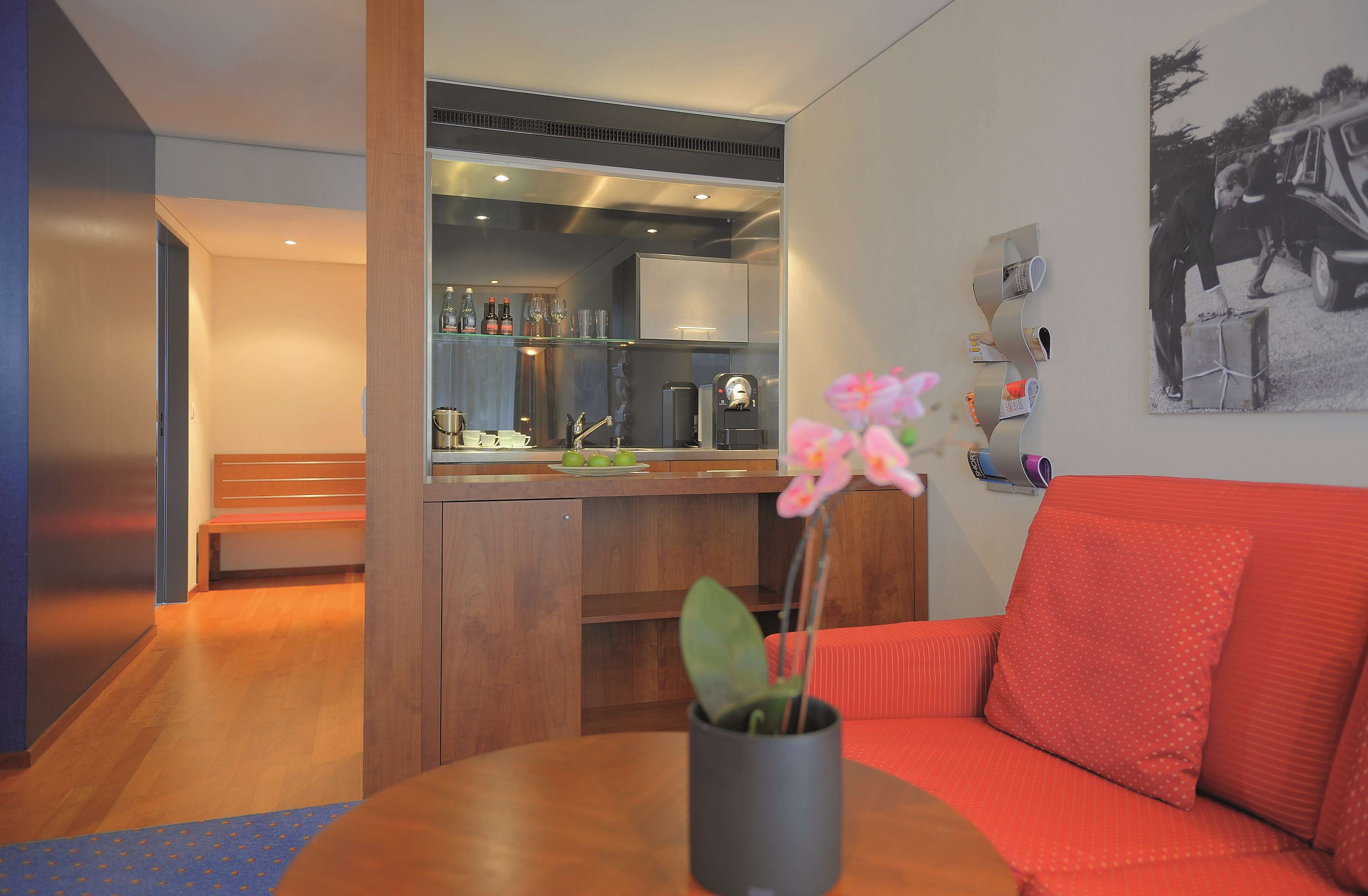 Uma sala de estar moderna com um sofá vermelho, uma mesa de centro com um vaso de flores, uma área de cozinha com balcão e um quadro pendurado na parede.