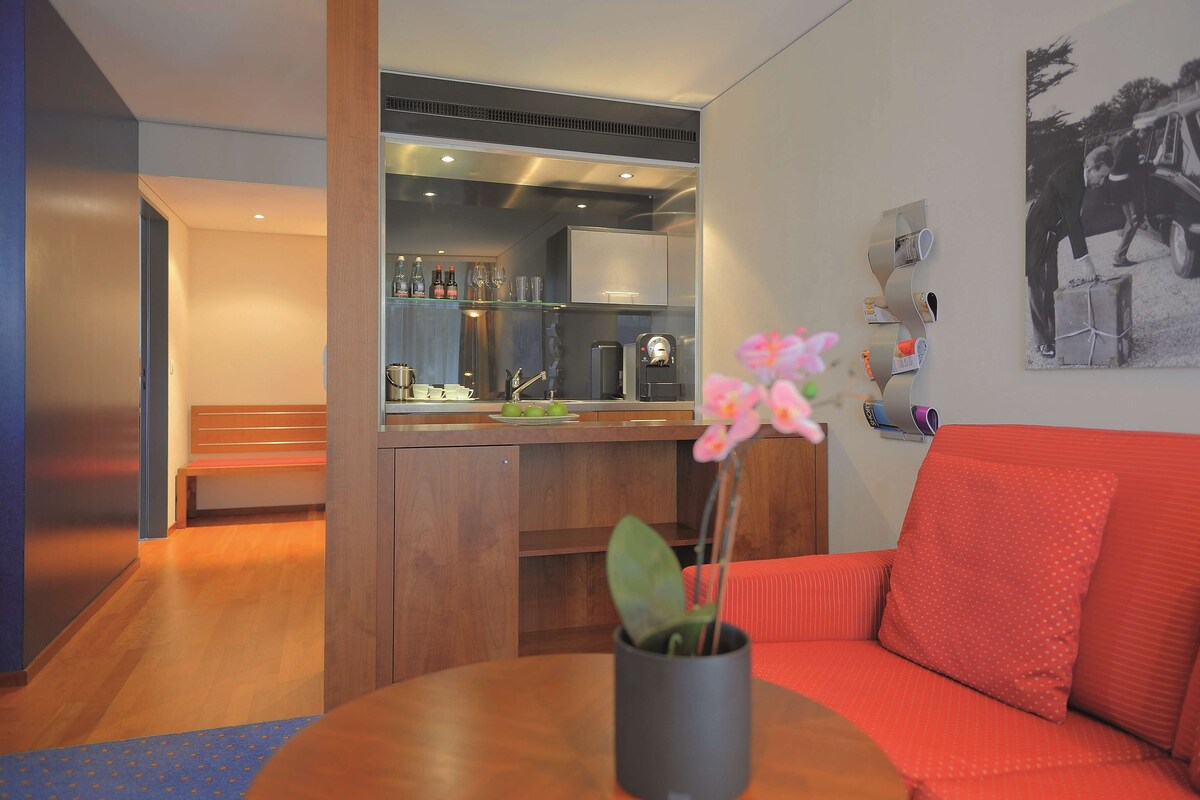 Una sala moderna con un sofá rojo, una mesa de centro con un florero, una zona de cocina con barra y un cuadro en la pared.