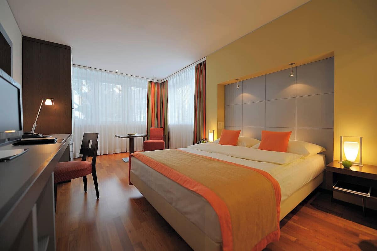 A hotel room with a large bed, a desk, a chair, a small table, and a nightstand with a lamp.