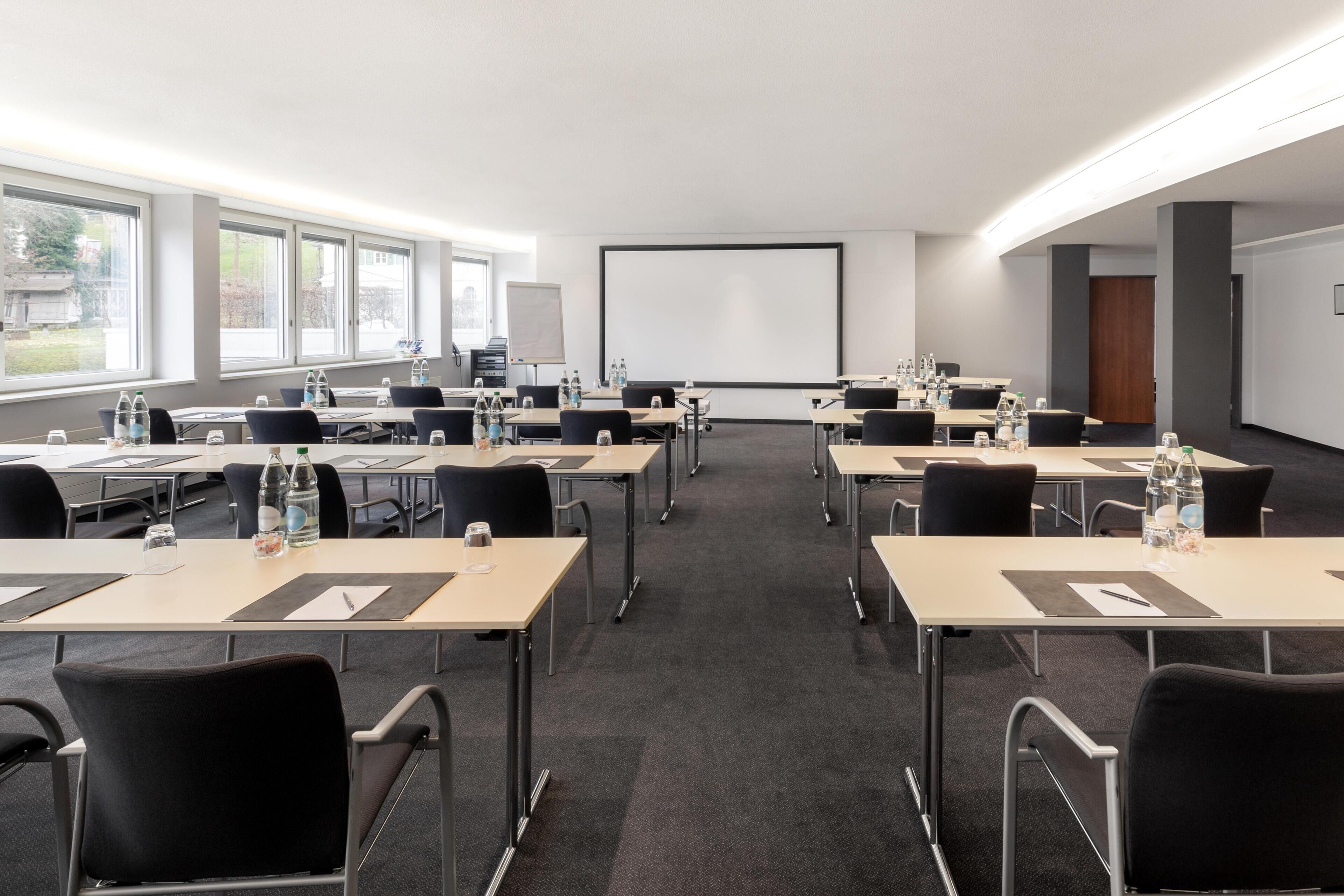 Un salón de conferencias con varias mesas, sillas, una pantalla de proyector y grandes ventanales.