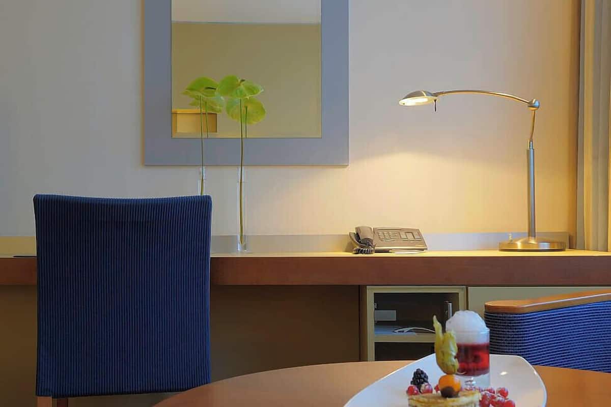 A hotel room with a round table, a blue chair, a desk with a lamp, a phone, and a plate with a dessert.