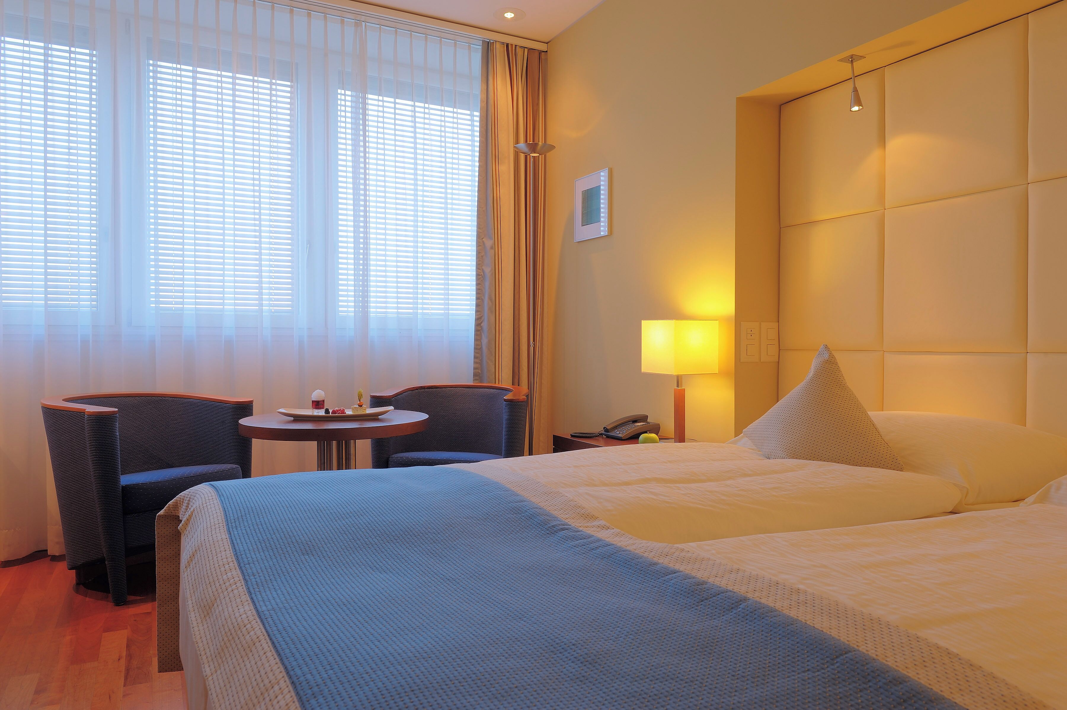 Ein Hotelzimmer mit zwei Betten, einem kleinen Tisch, einem Stuhl, einer Lampe und einem Fenster mit Vorhängen.