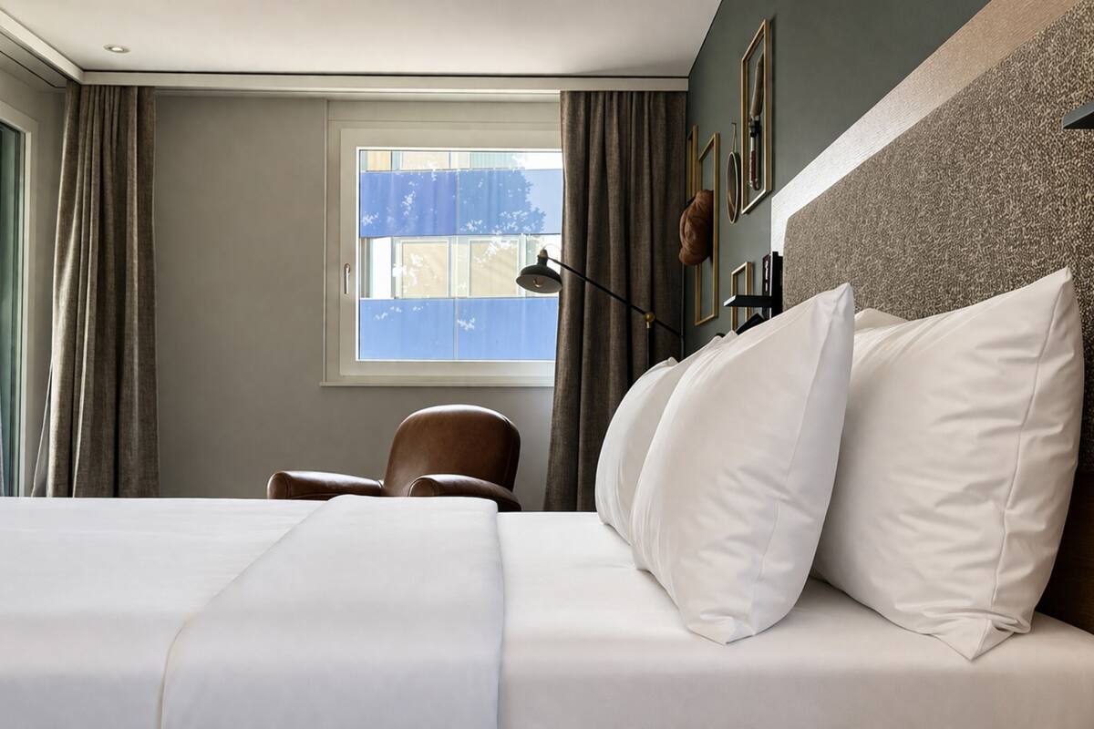A hotel room with a bed, a chair, a window with curtains, and a wall-mounted lamp.