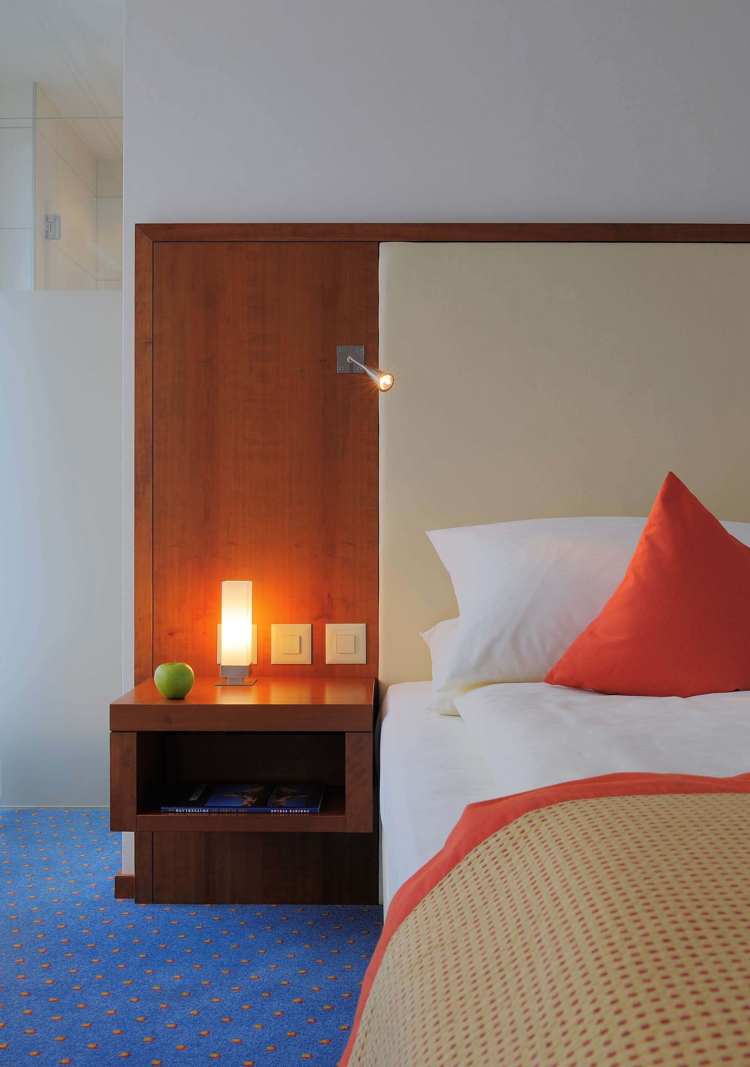 Ein Hotelzimmer mit Nachttisch aus Holz, einer Lampe, einem grünen Apfel und einem Bett mit weißen und roten Kissen.