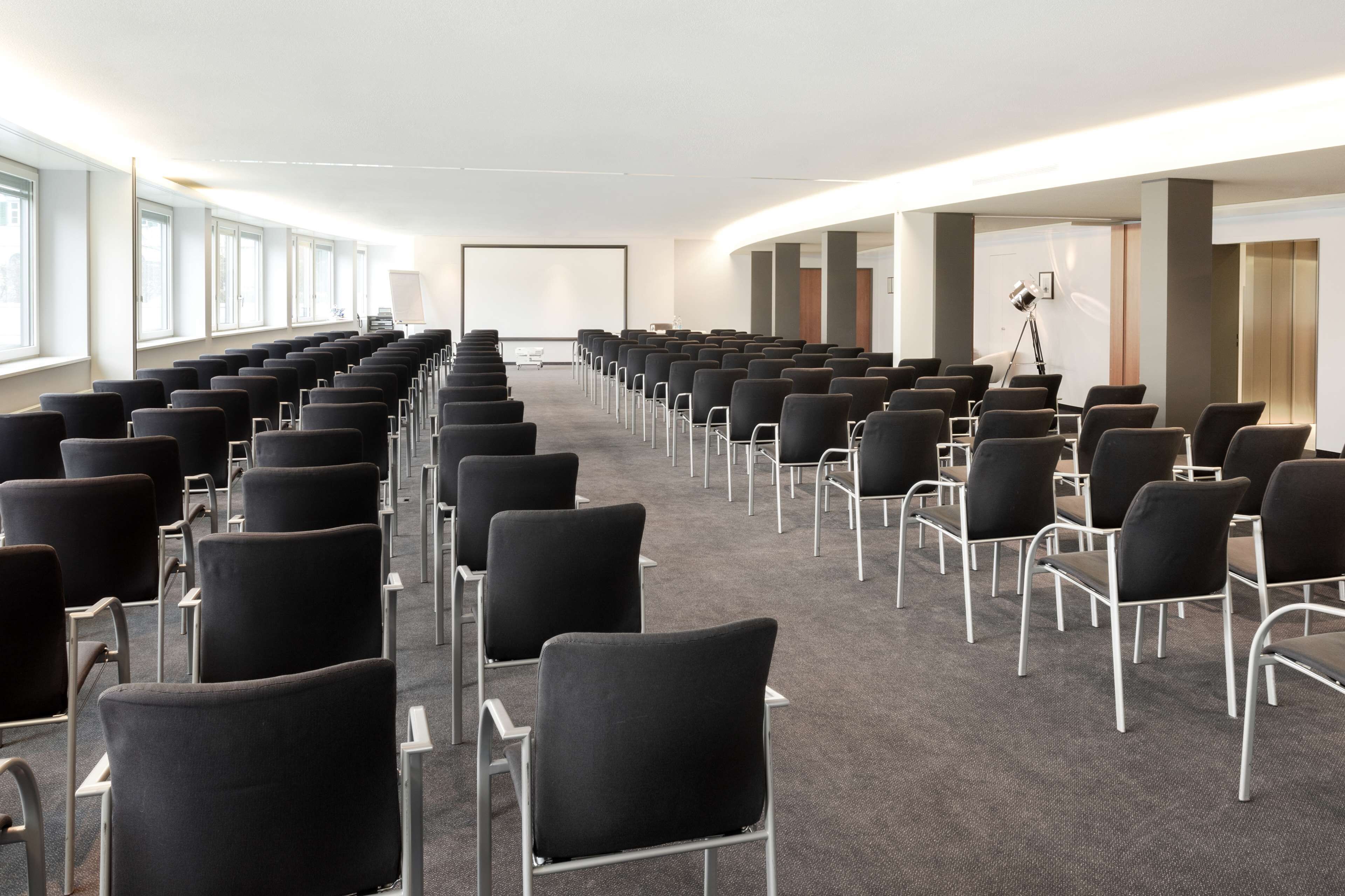 A conference room with rows of black chairs and a large screen at the front.