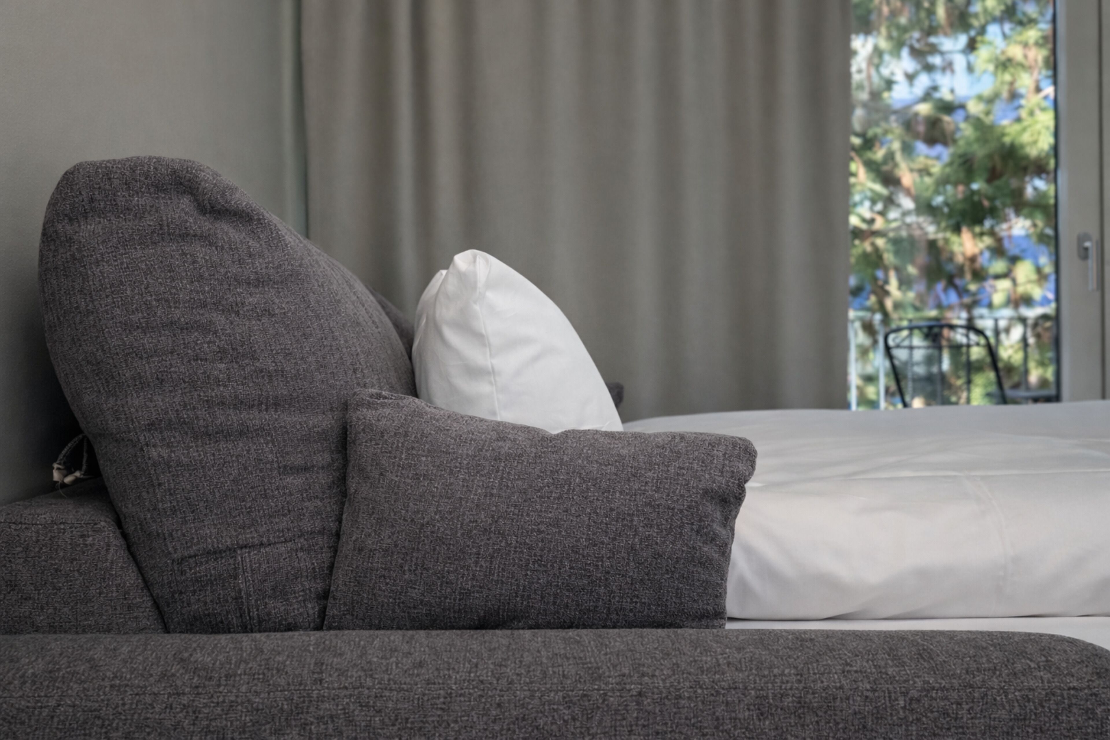Un lit avec une literie blanche, un canapé gris avec un coussin blanc, et une vue sur les arbres à travers une fenêtre.