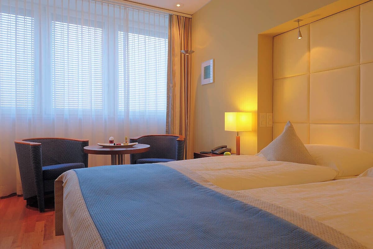 A hotel room with two beds, a small table with a plate of food, a chair, a lamp, and a window with curtains.