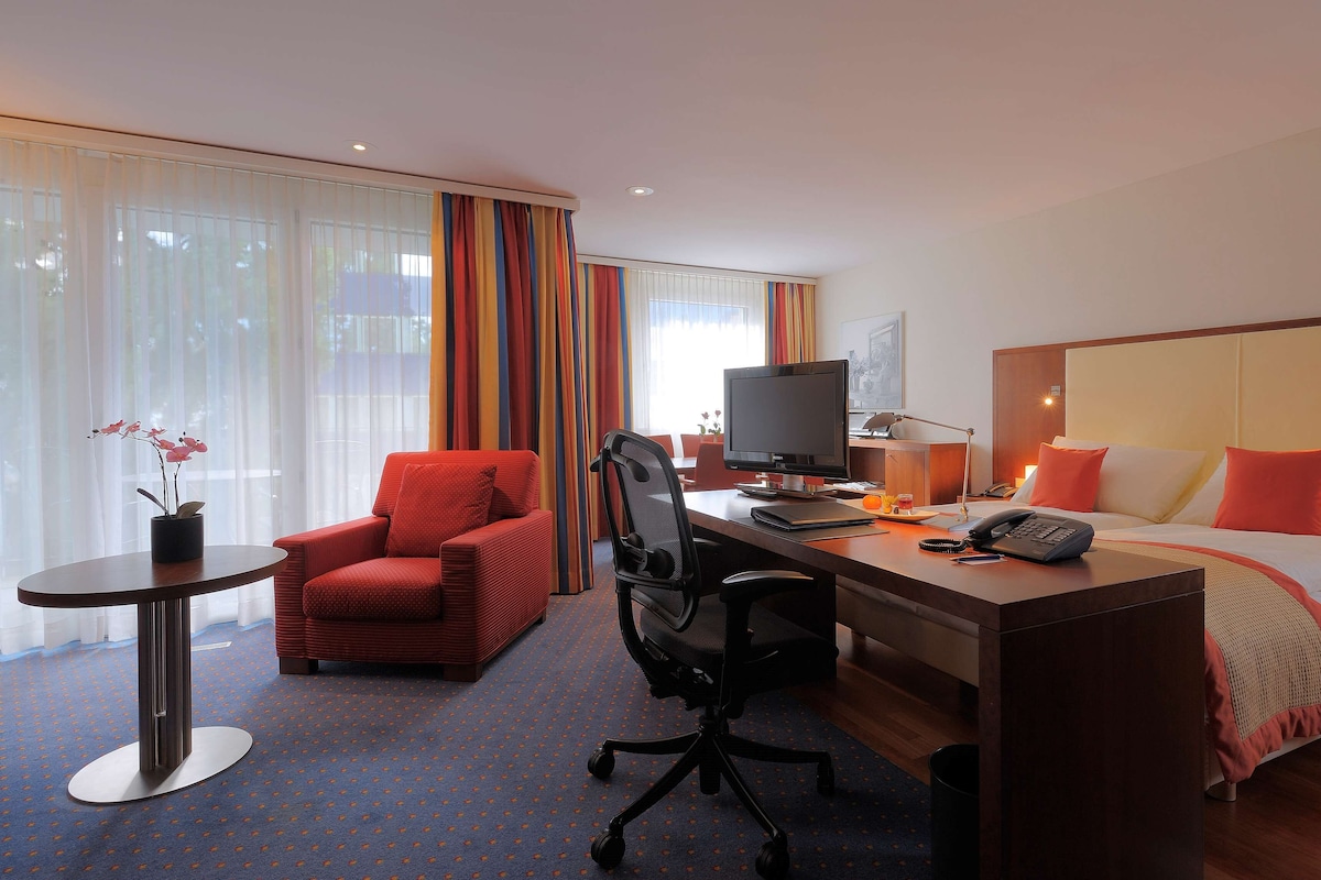 Habitación de hotel con cama, escritorio, silla, televisor y una pequeña mesa con un florero.