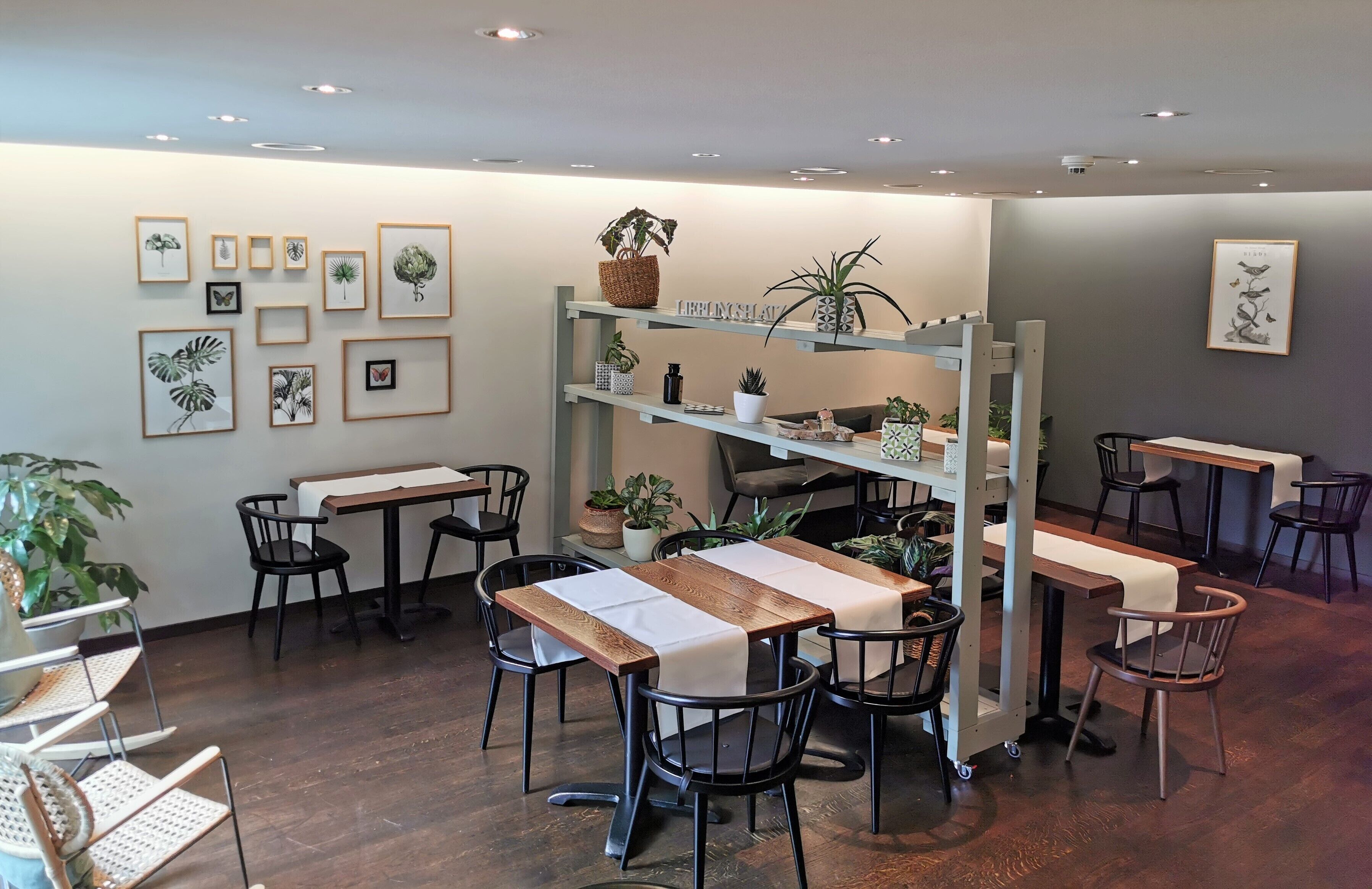 A café with wooden tables and chairs, framed botanical prints on the wall, and shelves with potted plants.