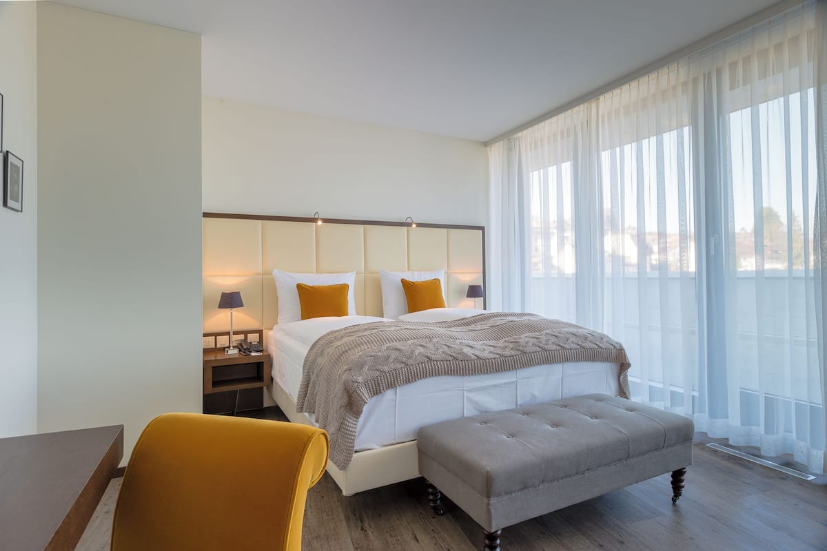A hotel room with a bed, a bedside table, a chair, and a large window with sheer curtains.