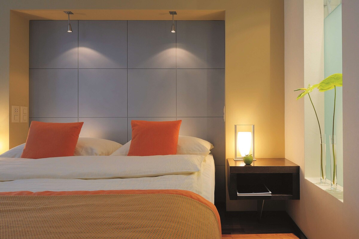 Una habitación de hotel moderna con una cama grande, almohadas anaranjadas, una mesita de noche con una lámpara y una ventana con una planta.