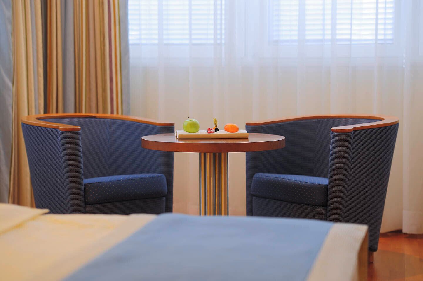 Una habitación de hotel con una cama, una mesita redonda con una bandeja de frutas y dos sillones tapizados en azul.