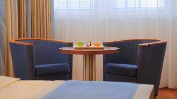 Una habitación de hotel con una cama, una mesita redonda con una bandeja de frutas y dos sillones tapizados en azul.