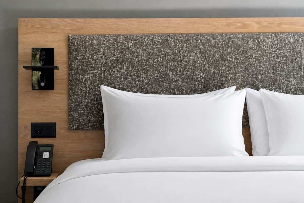 A hotel room with a wooden headboard, a textured wall panel, a bedside telephone, and white bedding.