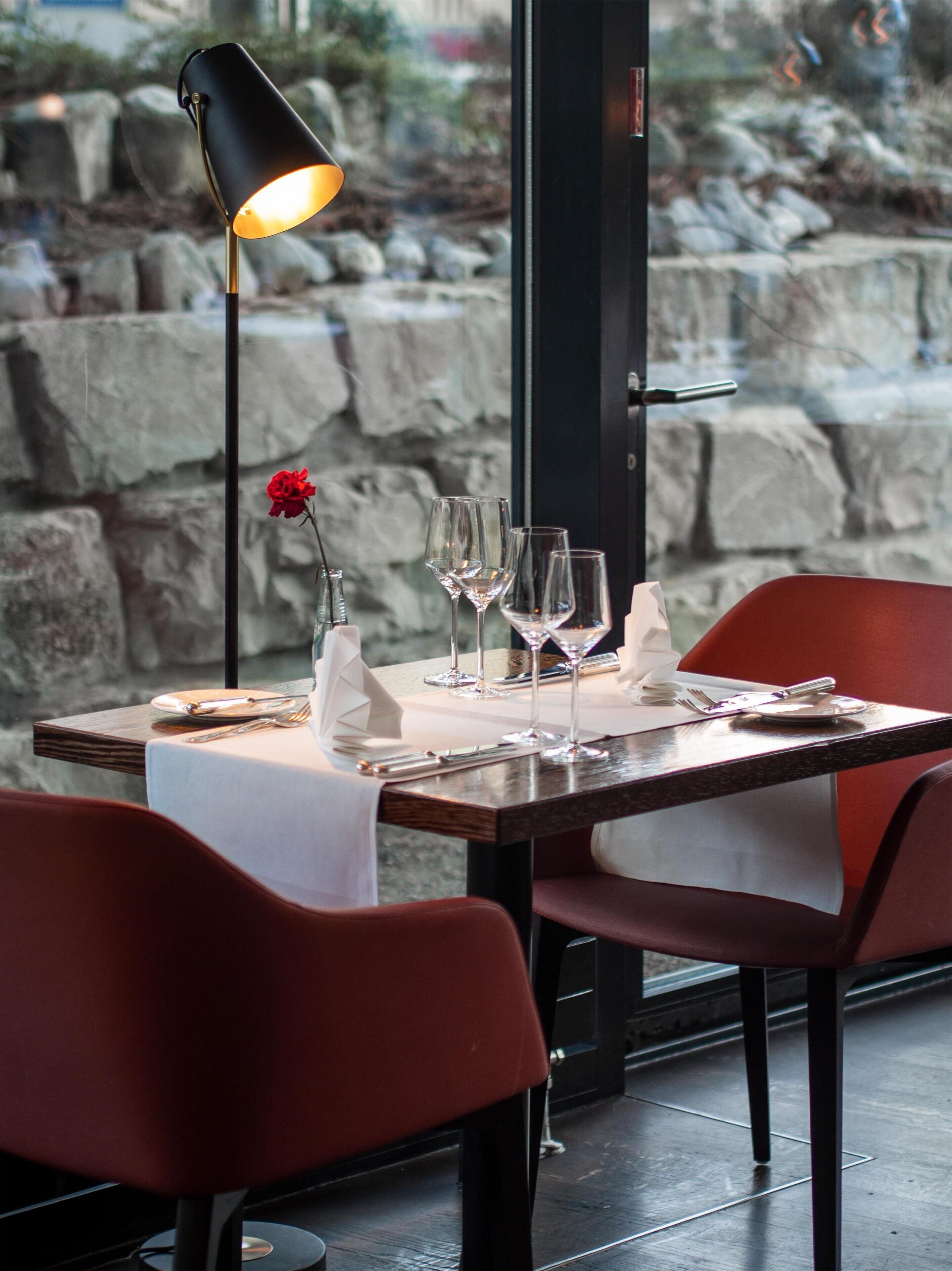 Une table à manger dressée avec des verres à vin, une rose rouge et une bougie, le tout sur fond d’un mur en pierre et d’une lampe.
