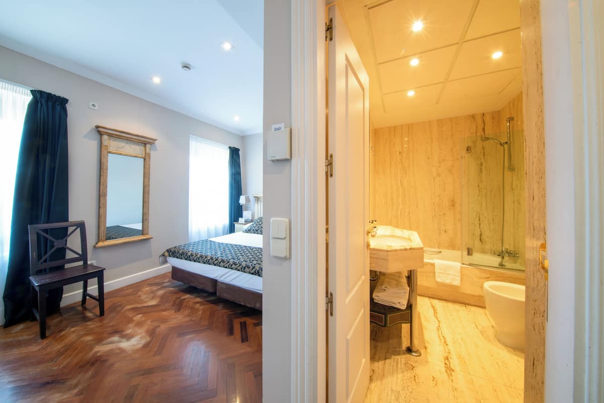 Un dormitorio con cama, una silla y una ventana con cortinas. El suelo es de madera y hay un espejo en la pared.