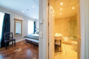 Ein Schlafzimmer mit einem Bett, einem Sessel und einem Fenster mit Vorhängen. Der Raum hat Holzböden und einen Spiegel an der Wand.
