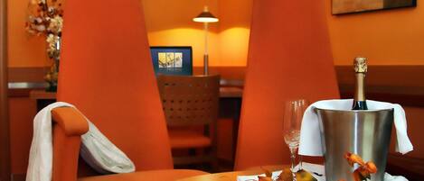 A room with orange walls, a round table, two orange chairs, a metal bucket with a bottle, a glass, and a vase with dried flowers.