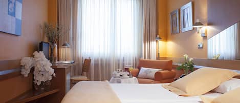 A hotel room with a large bed, a sofa, a desk with a lamp, and a window with curtains.