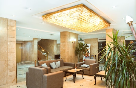 A spacious lobby with a high ceiling, a large chandelier, comfortable seating, and a staircase leading to an upper floor.