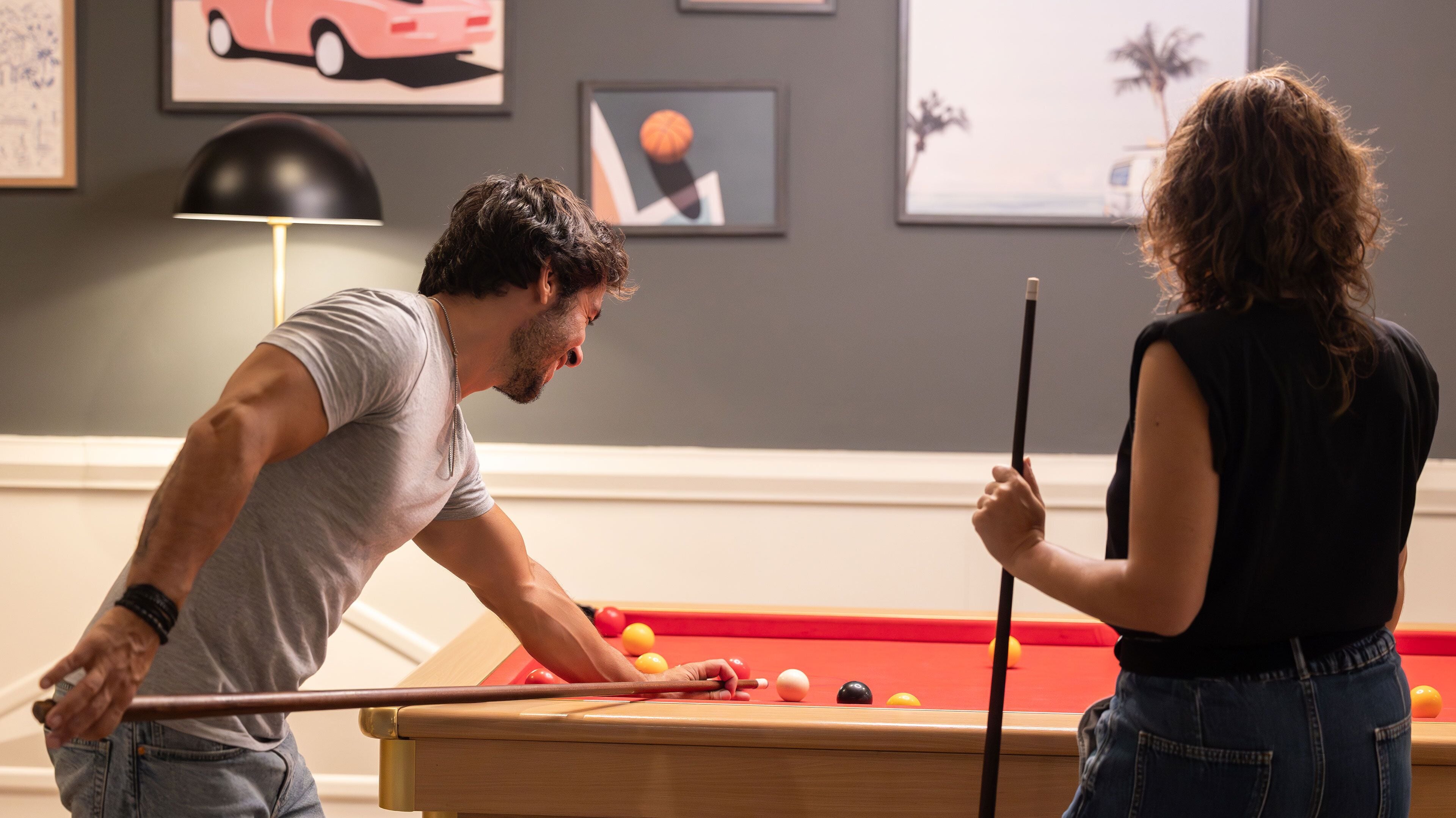 Zwei Personen spielen Billard in einem Raum mit einer Billardtisch, Bildern an den Wänden und einer Lampe.