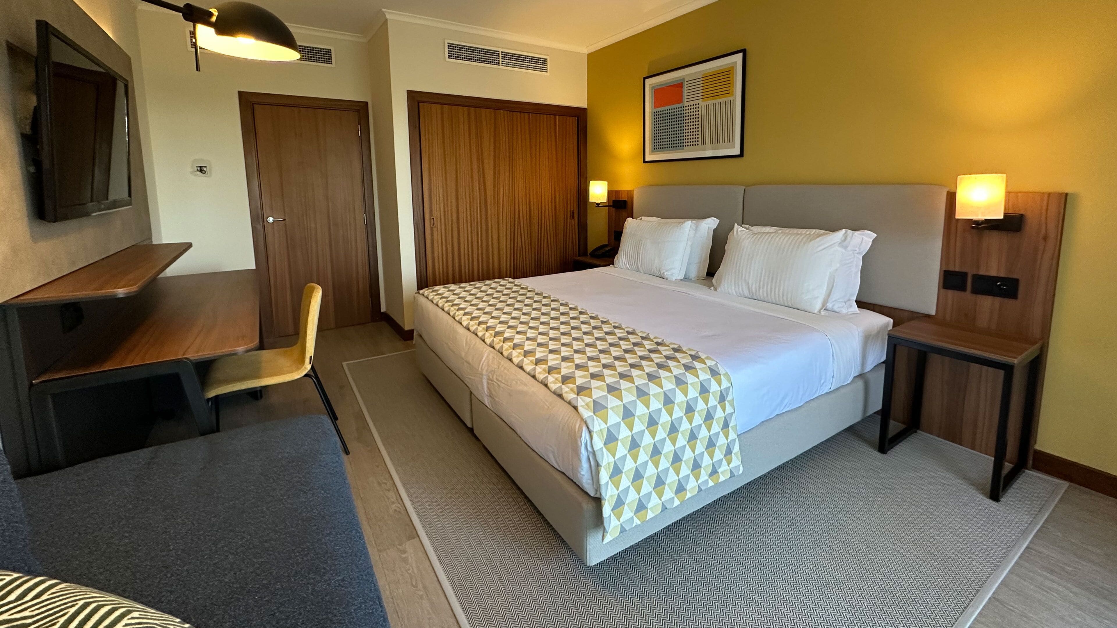 A hotel room with a bed, a desk, a chair, a wardrobe, and a bedside table with a lamp.