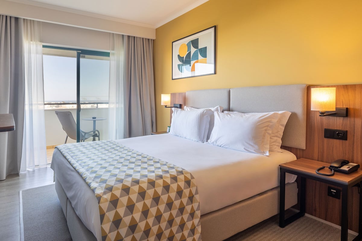 Una habitación de hotel con cama, escritorio, silla, una mesita y ventana con cortinas.