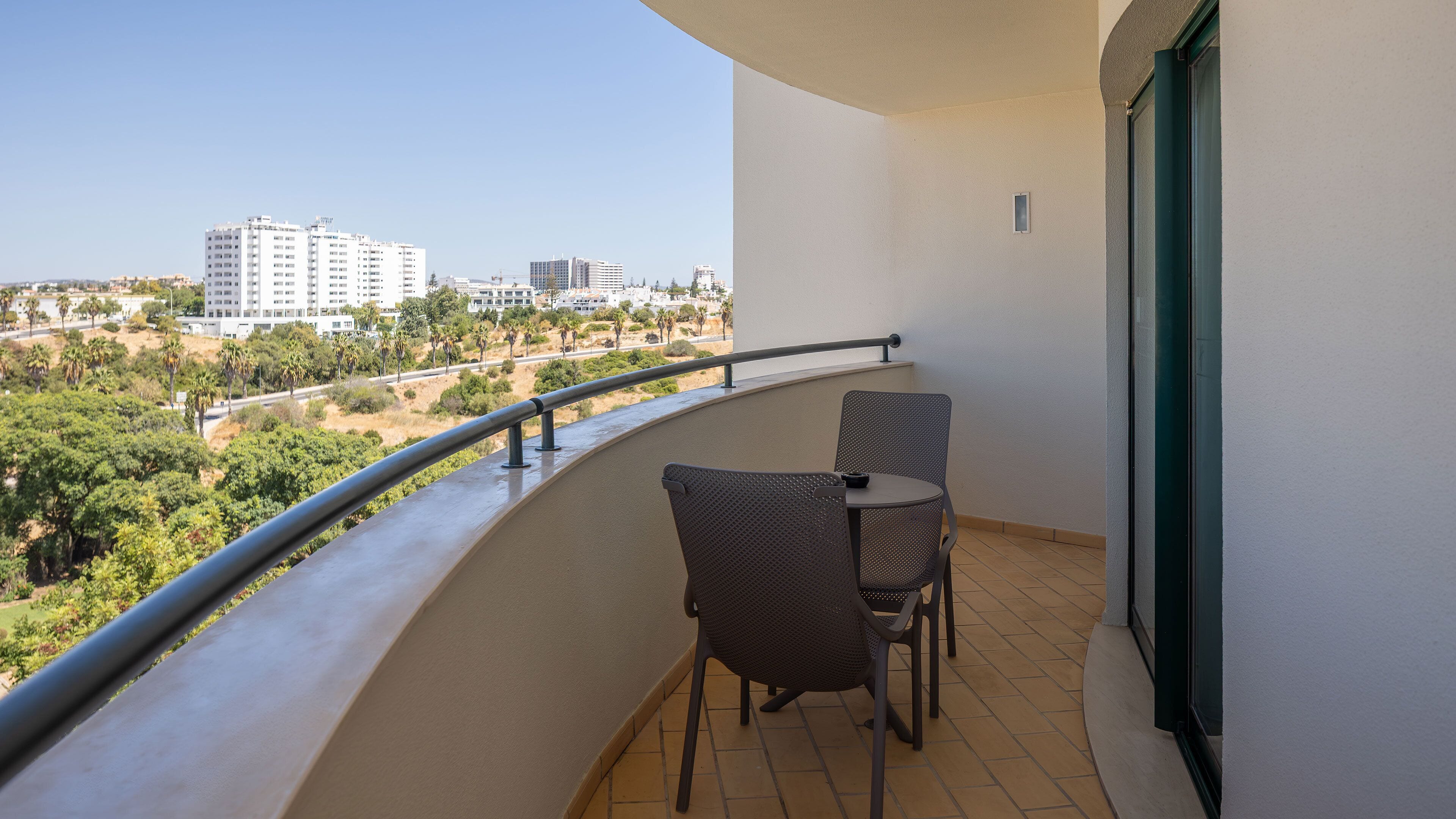 Un balcon avec deux chaises et une table, donnant sur un paysage urbain composé d’immeubles et d’espaces verts.