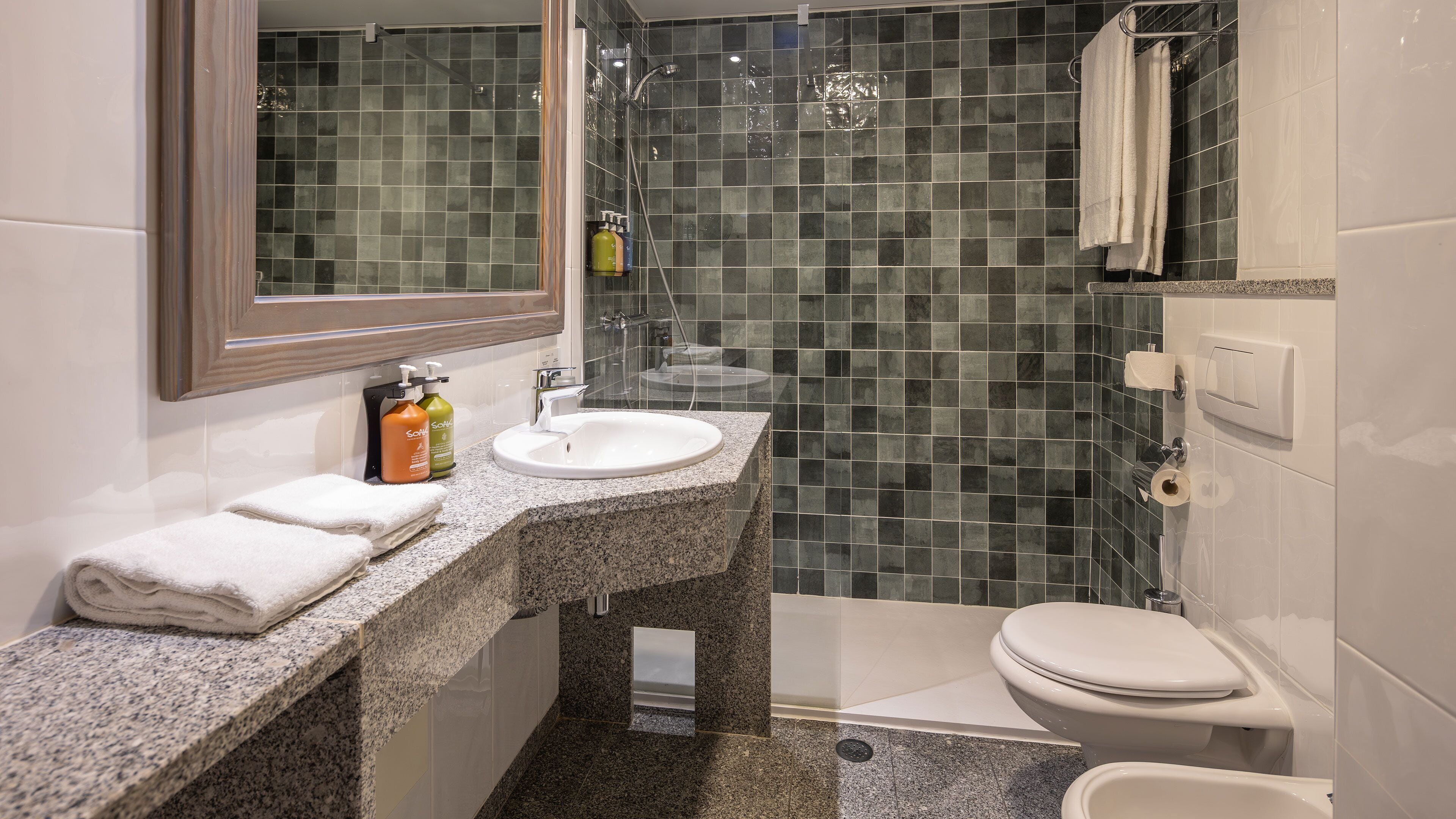 Un baño con encimera de granito, un lavabo blanco, un espejo, cabina de ducha con mampara de vidrio y un inodoro.