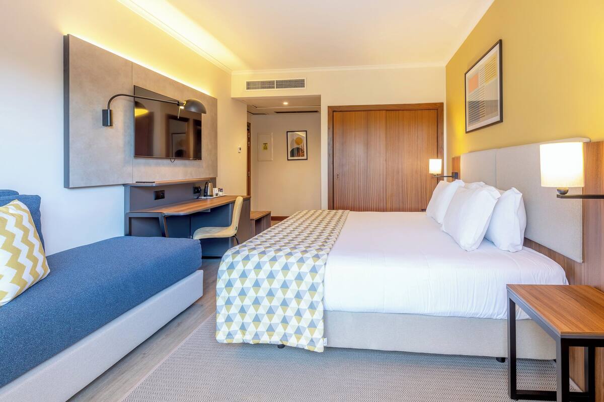Una habitación de hotel con una cama, un escritorio, una silla, un espejo y una mesita de noche.