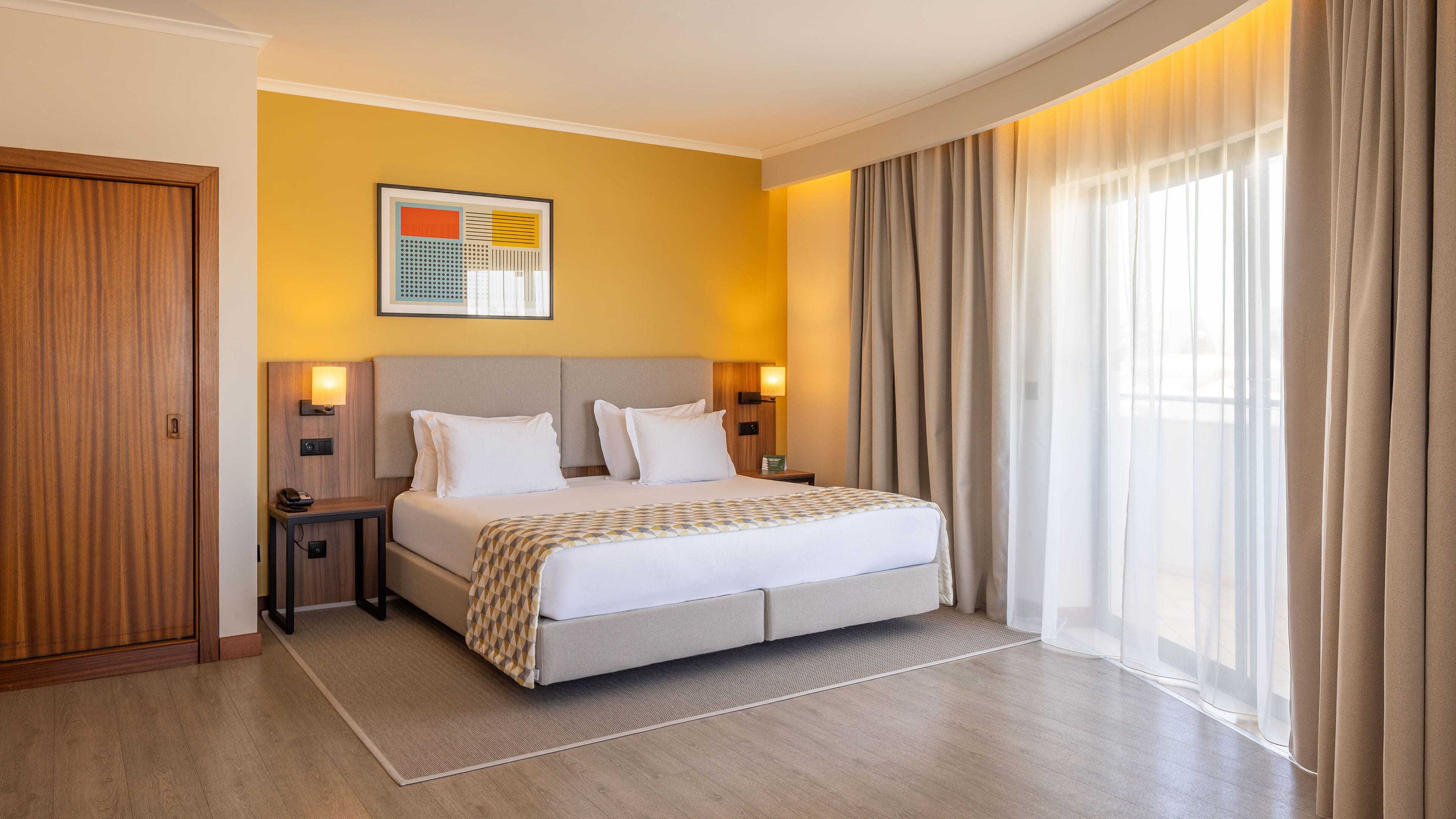 Habitación de hotel con cama, mesitas de noche, una obra de arte en la pared y un amplio ventanal con cortinas.