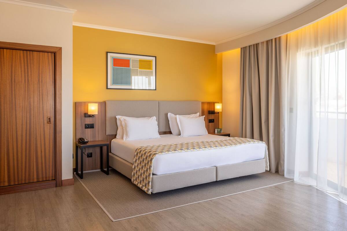 Habitación de hotel con cama, mesitas de noche, una obra de arte en la pared y un amplio ventanal con cortinas.