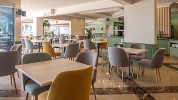 Een ruime eethoek met meerdere tafels en stoelen, een keuken in de achtergrond en een groot raam dat voor natuurlijk licht zorgt.