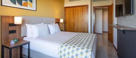 A hotel room with a large bed, a bedside table with a lamp, a wooden headboard, a wall-mounted TV, and a door leading to another room.
