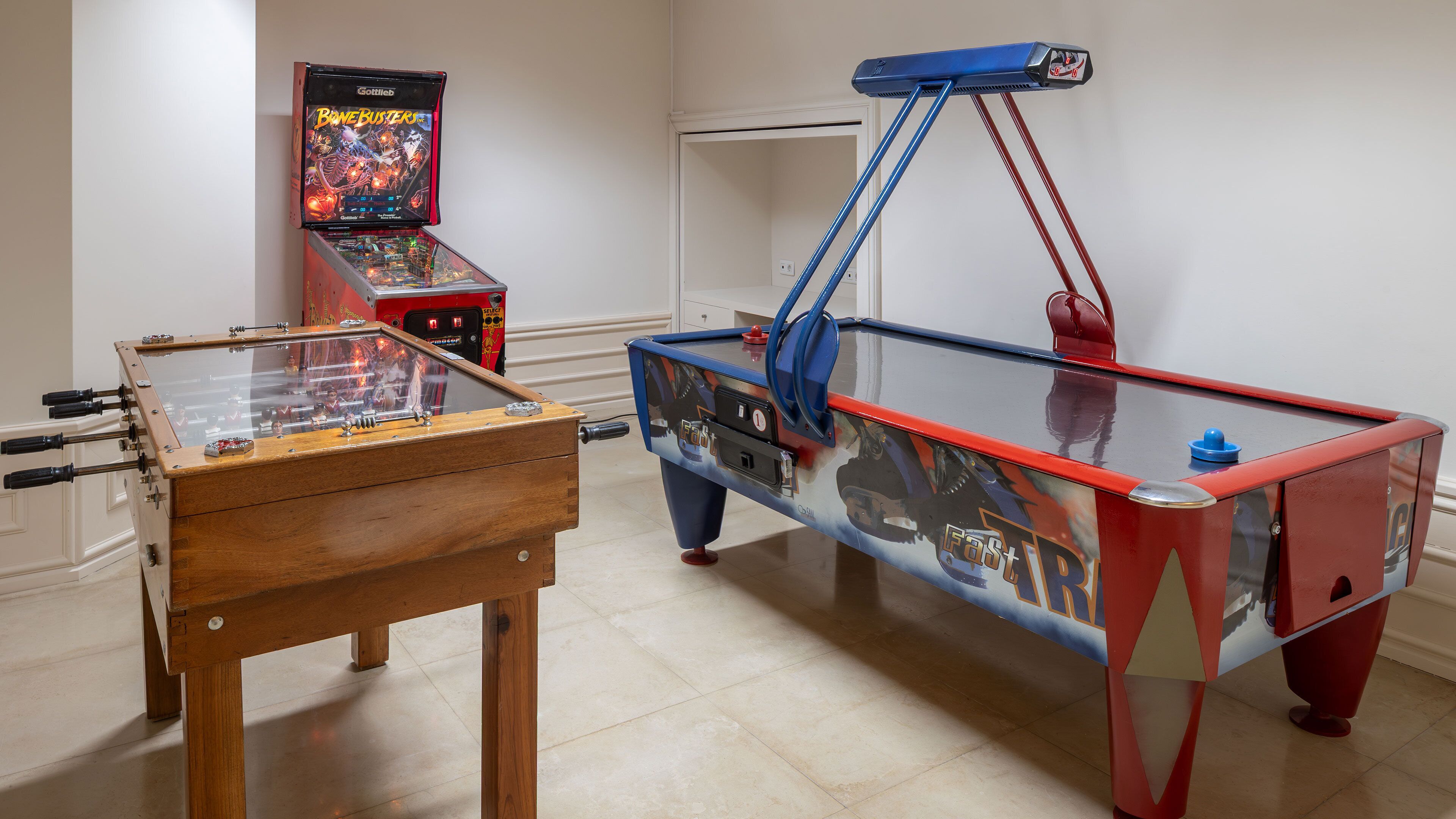 A room with two foosball tables, one wooden and one with a red and blue design, and a vintage arcade machine.