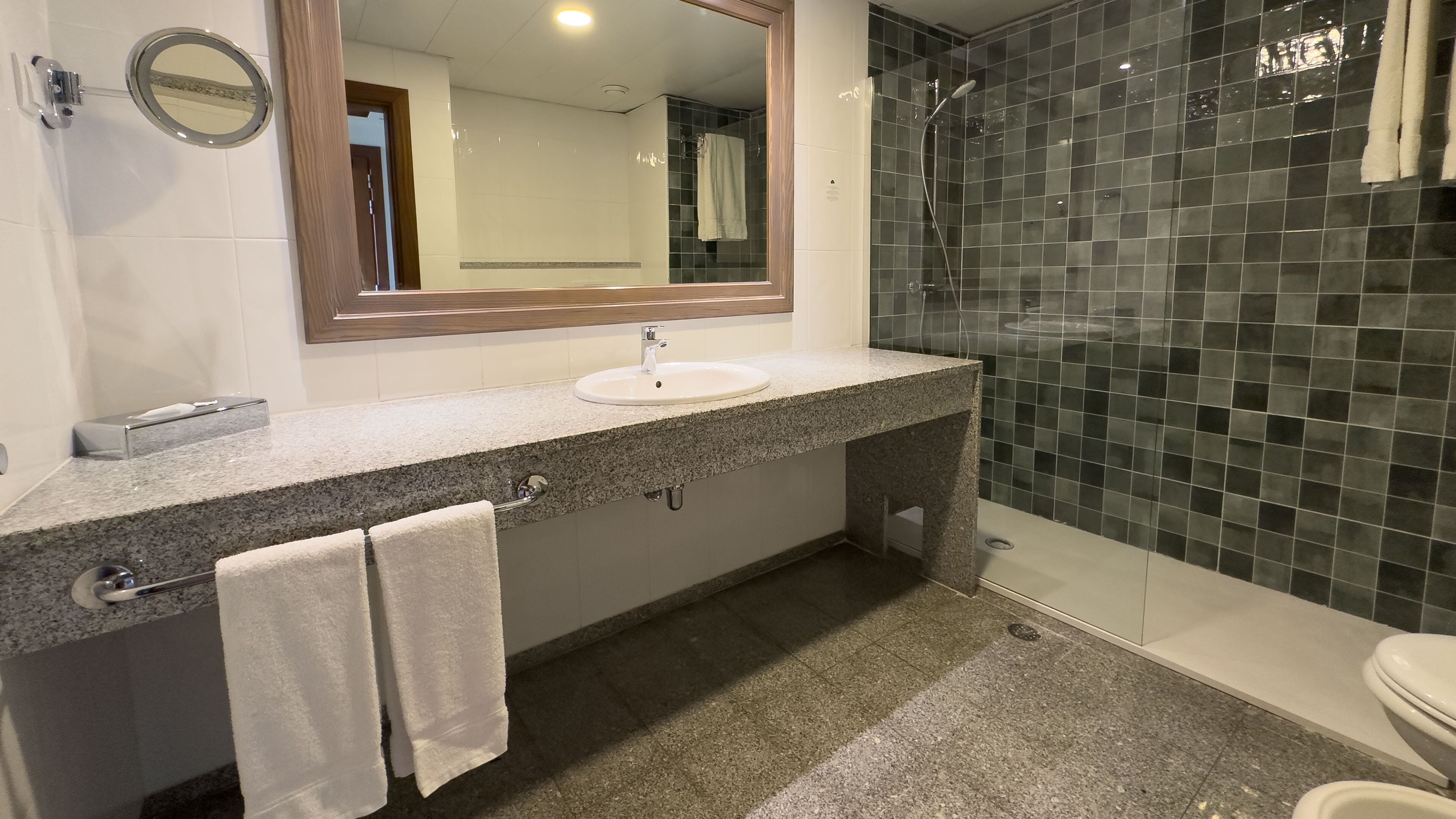 Un baño con encimera de granito, lavamanos, espejo, toallero y zona de ducha.