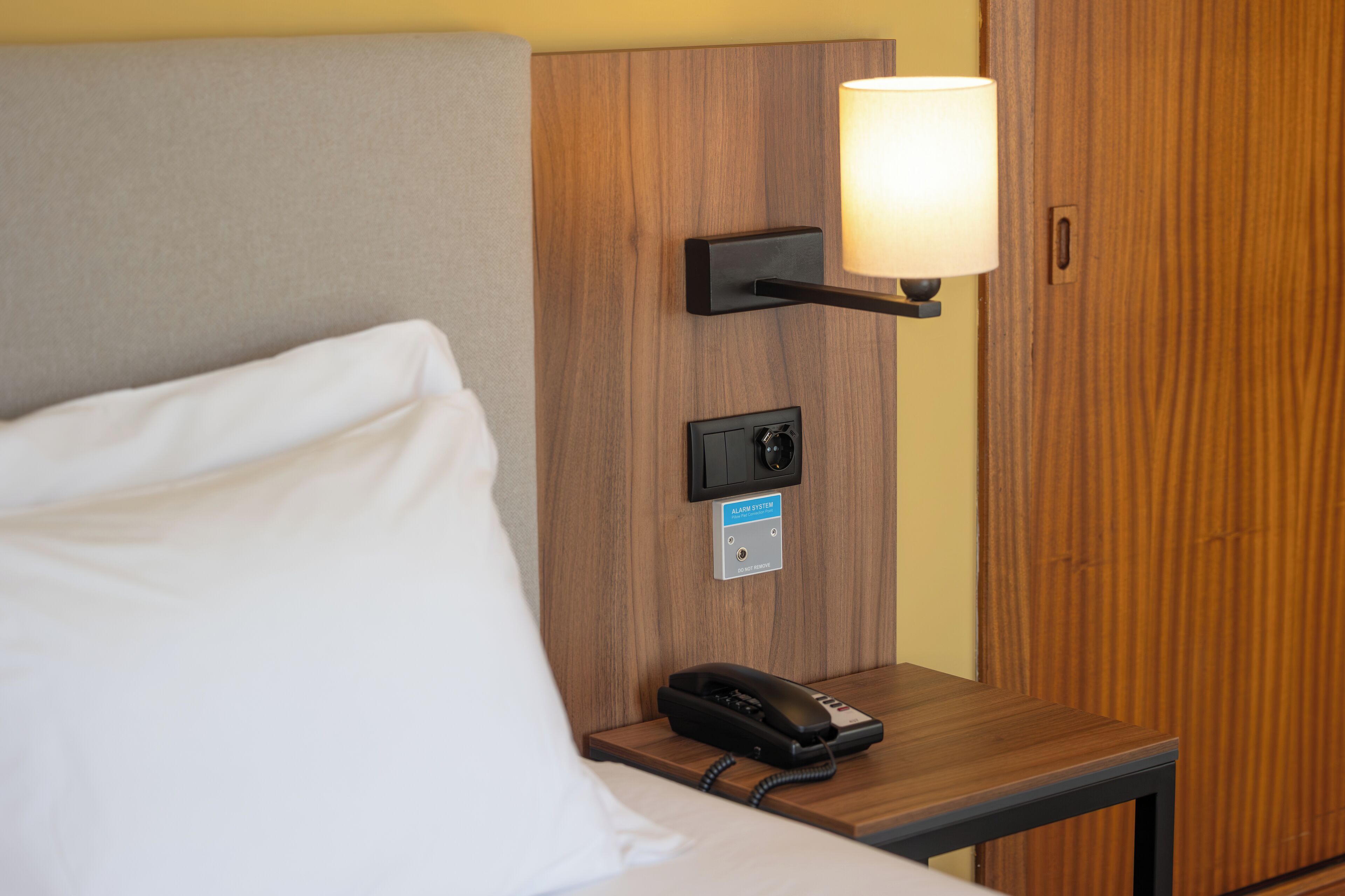 Una habitación de hotel con una cama, una mesita de noche con lámpara, un reloj despertador y un teléfono.