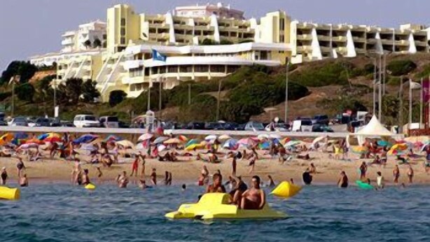 Ein Strand mit Menschen auf Wassersportgeräten und Sonnenschirmen, ein großes Gebäude im Hintergrund und ein klarer Himmel.
