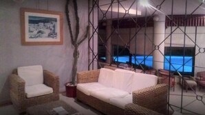 Ein Zimmer mit einem Korbssessel, einem Sessel, einer Topfpflanze und Blick auf einen Pool durch eine fensterartige geometrische Struktur.