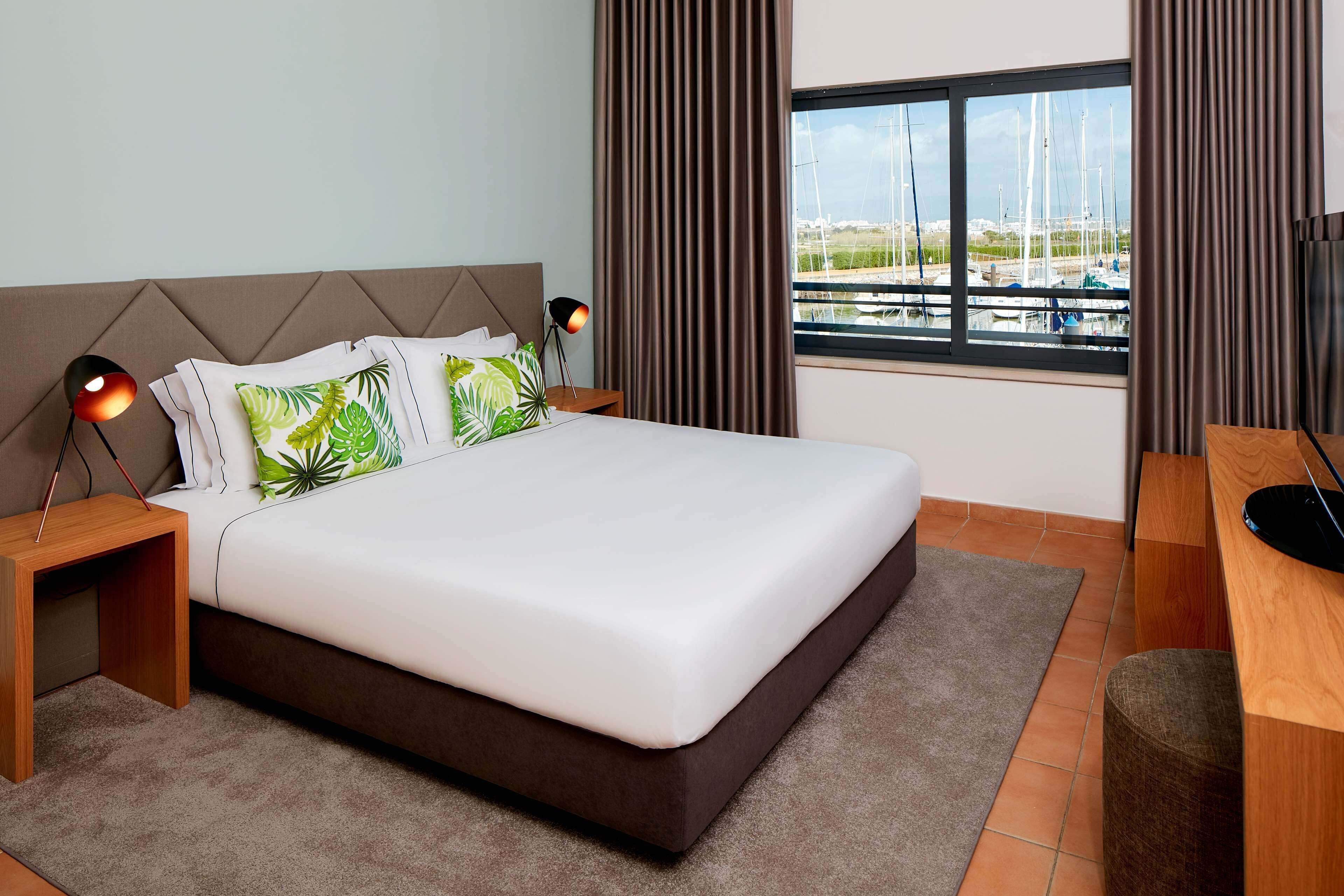 Een moderne hotelkamer met een groot bed, twee nachtkastjes, een bureau en uitzicht op boten door een raam.