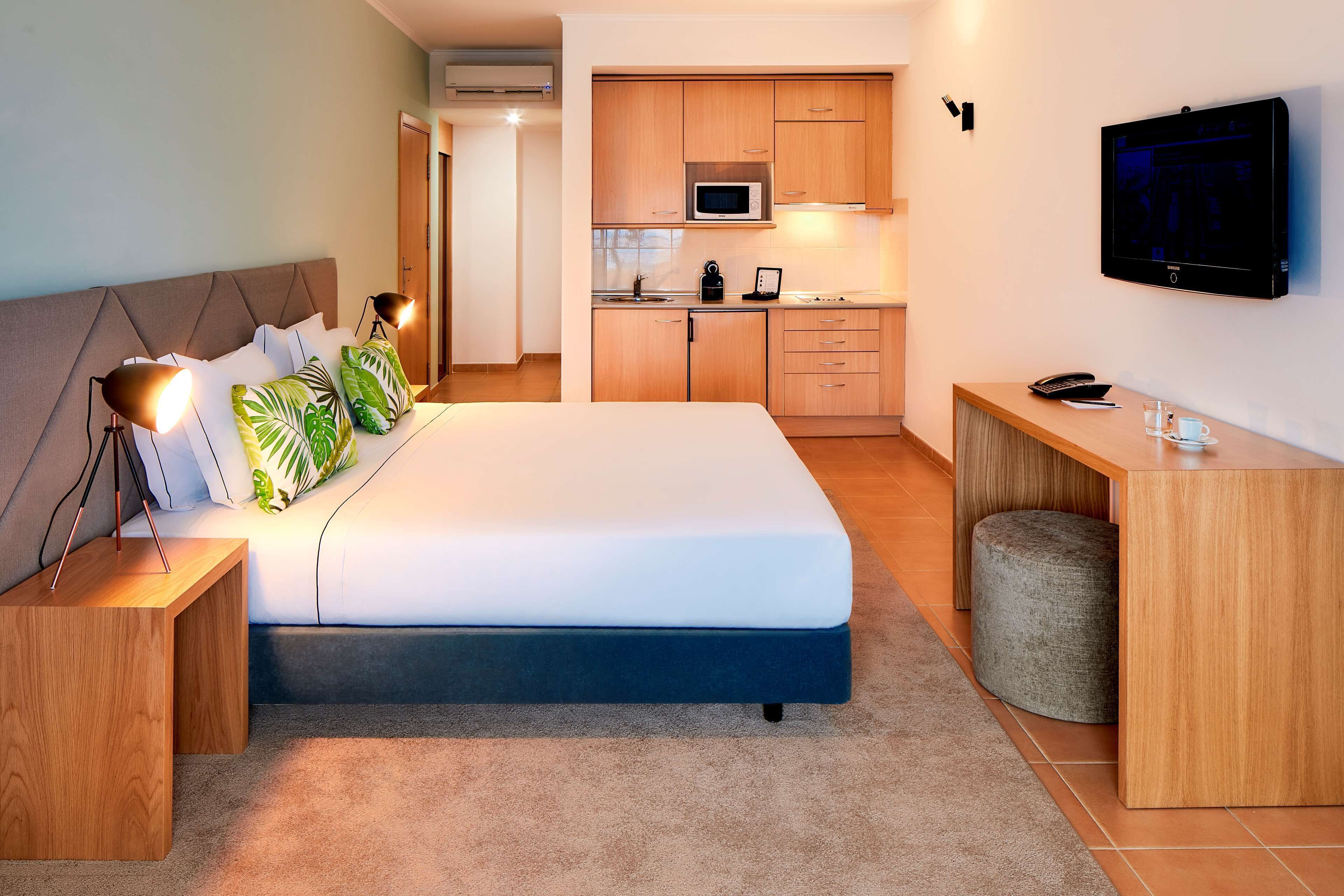 A modern hotel room with a large bed, bedside lamps, a desk, a television, and a kitchenette.