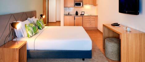 Quarto de hotel moderno com uma cama grande, abajures, uma escrivaninha, televisão e uma cozinha compacta.