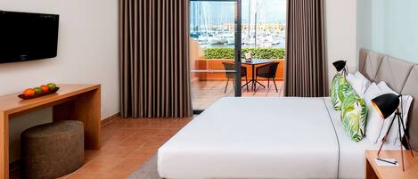 Habitación de hotel moderna con una cama grande, un escritorio con computadora, televisor y balcón con vista a barcos y un puerto deportivo.