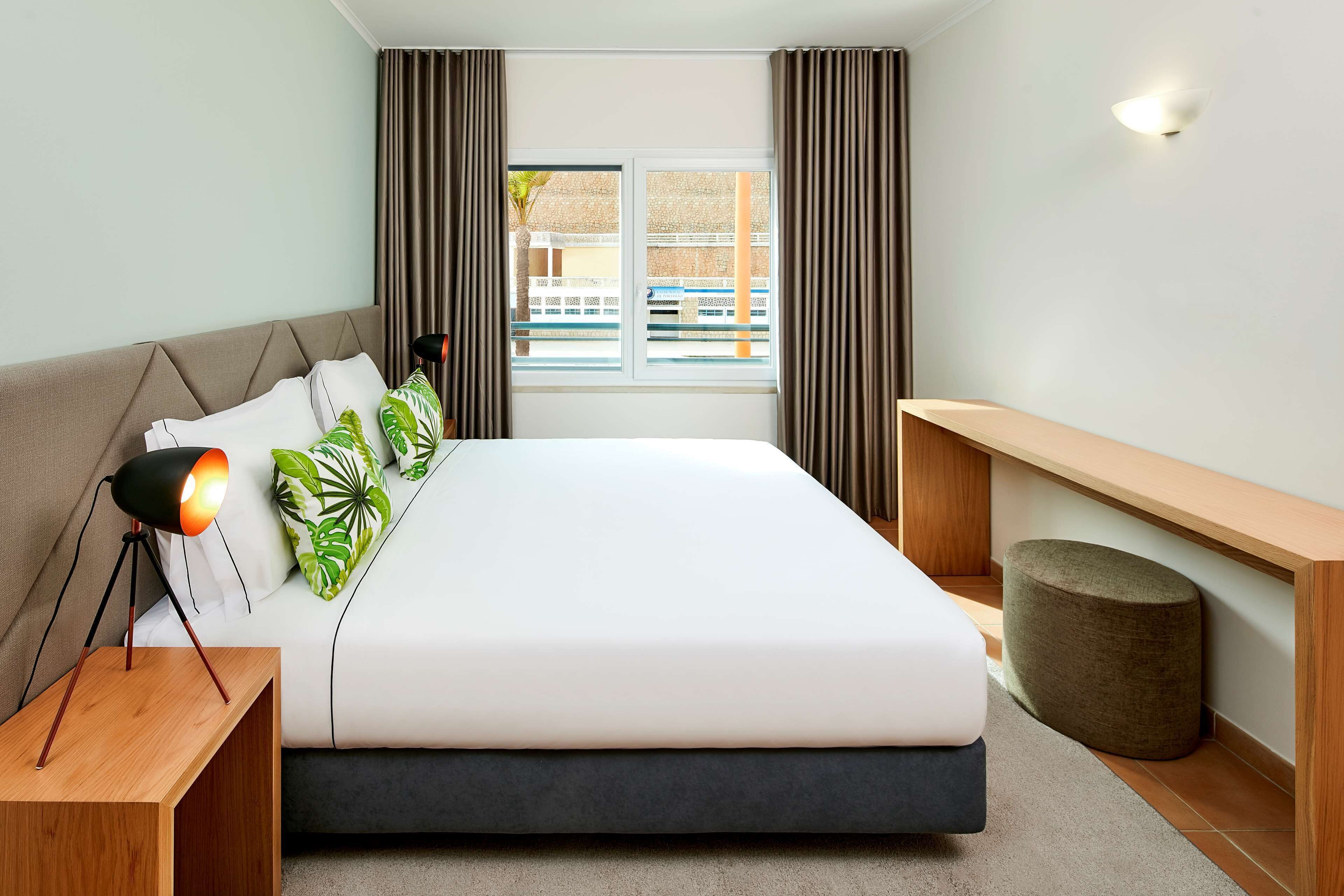 A modern bedroom with a bed, bedside lamp, and a view of the outside.