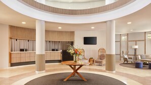 A spacious lobby with a central round table, wooden paneling, a flat-screen TV, and a seating area with a sofa and chairs.