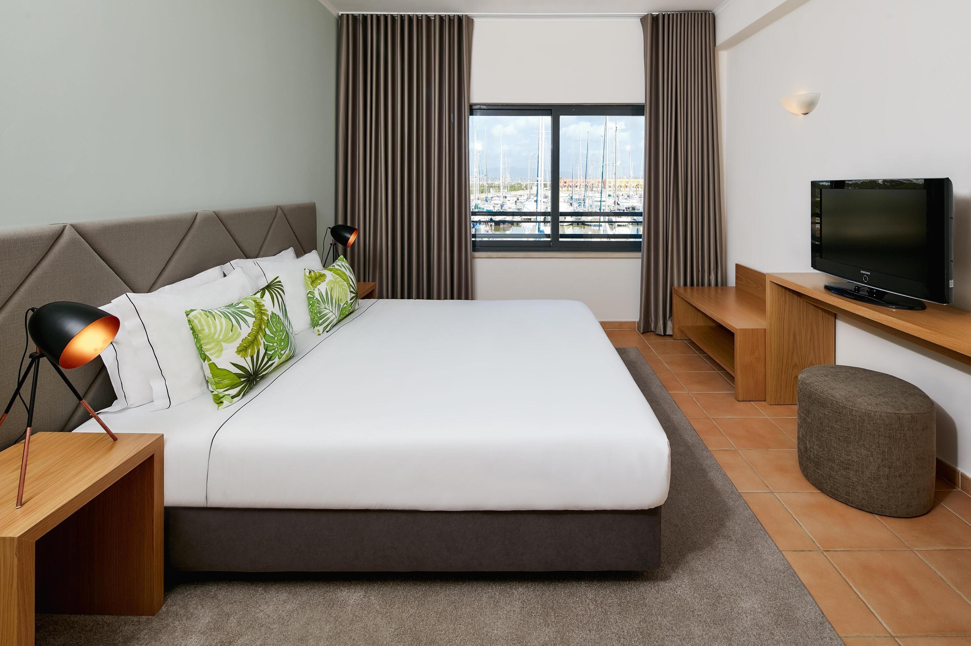 A modern hotel room with a large bed, a TV, a desk, and a view of boats through the window.