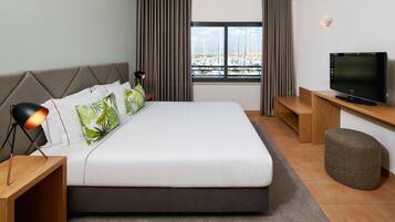 Moderni hotellihuone, jossa on suuri sänky, televisio, työpöytä ja ikkunasta avautuva näkymä veneisiin.