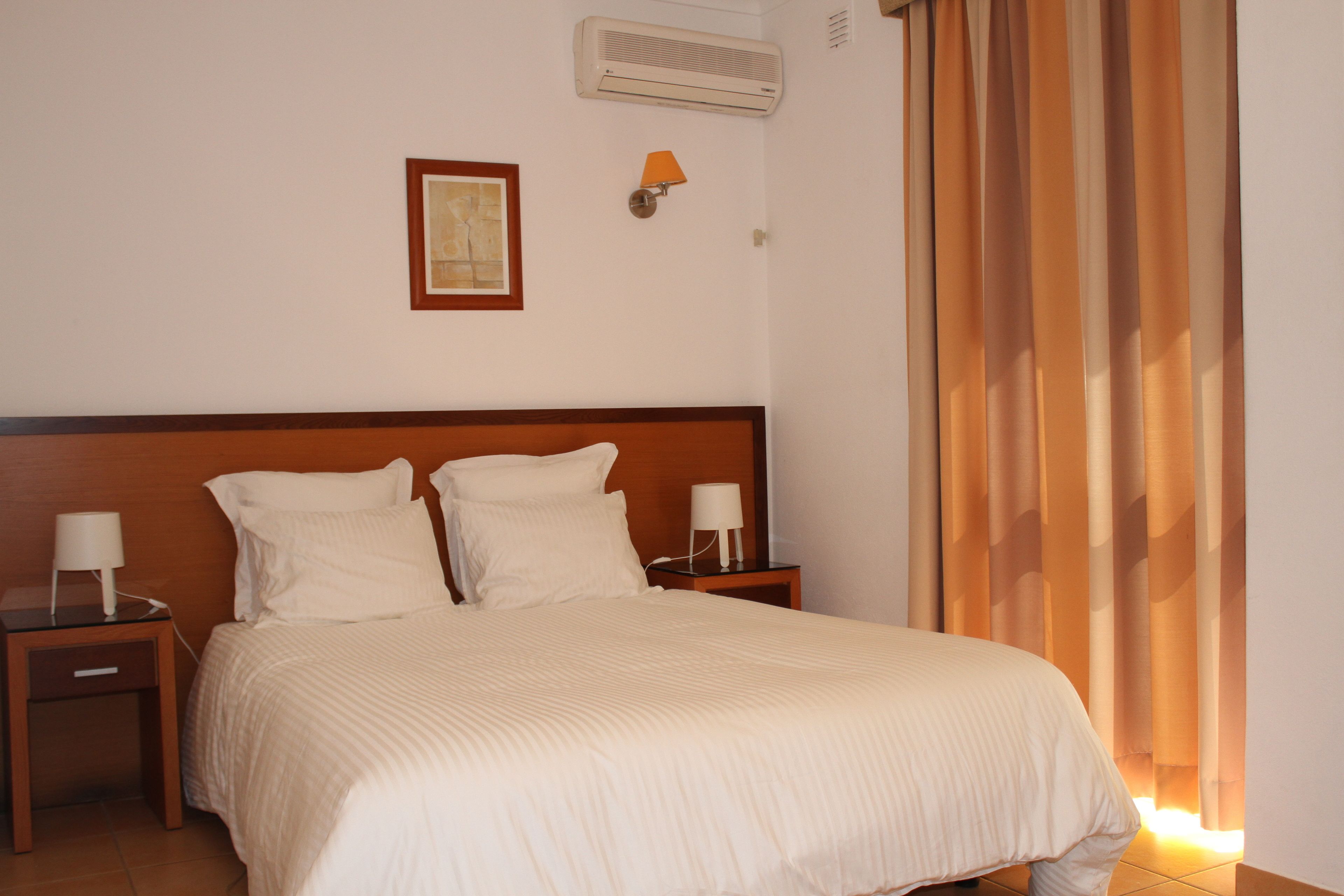 Een hotelkamer met een groot bed, twee nachtkastjes met lampen, een wandairconditioner, een inlijsting afbeelding en een raam met gordijnen.