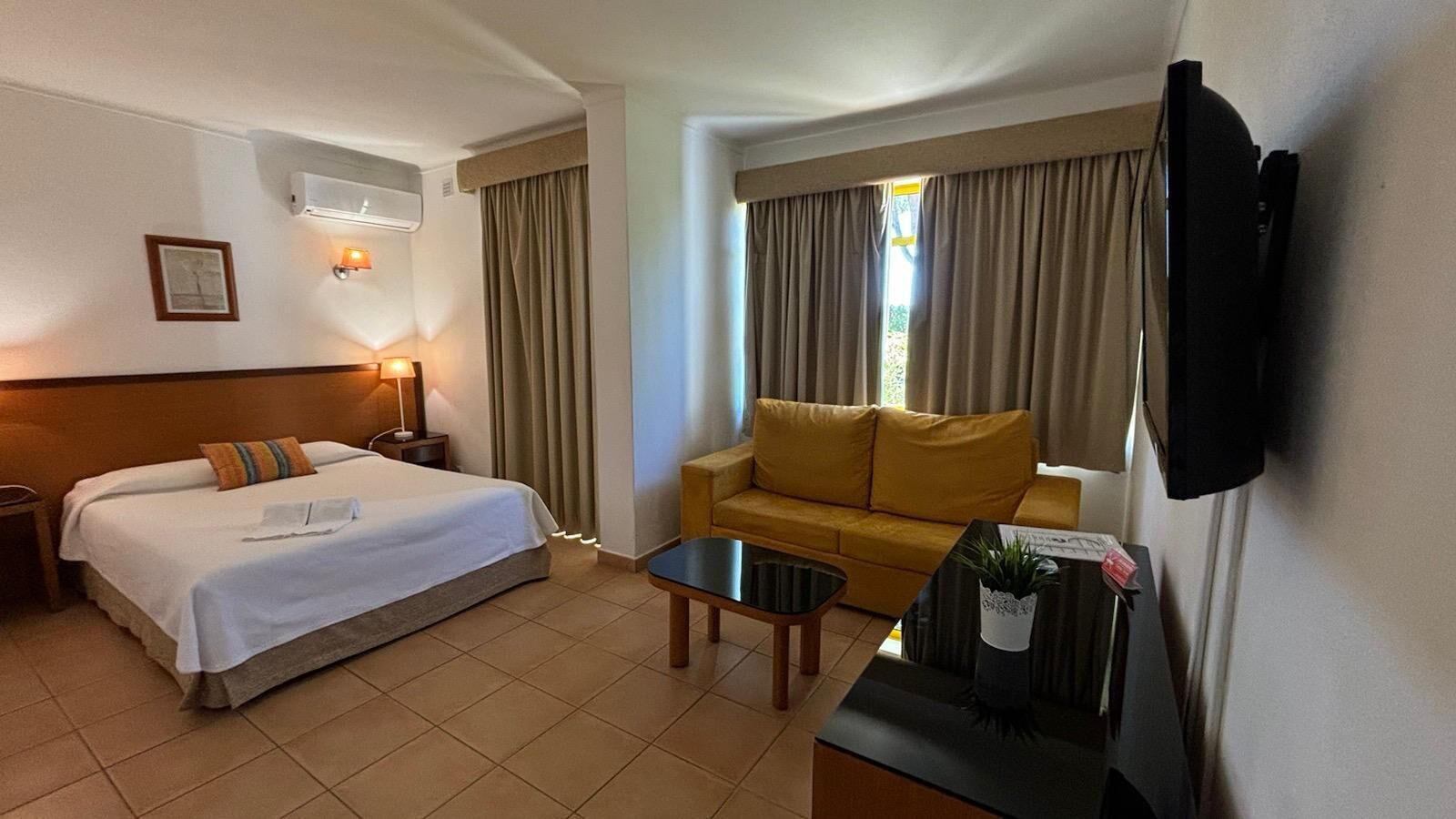 A hotel room with a bed, a sofa, a coffee table, a TV, and a window with curtains.